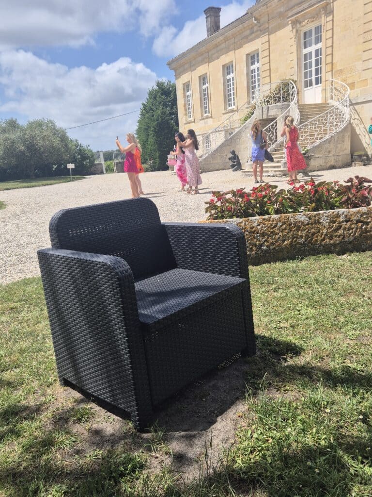 Fauteuil noir en résine tressée devant un château en pierre avec des touristes par temps ensoleillé.